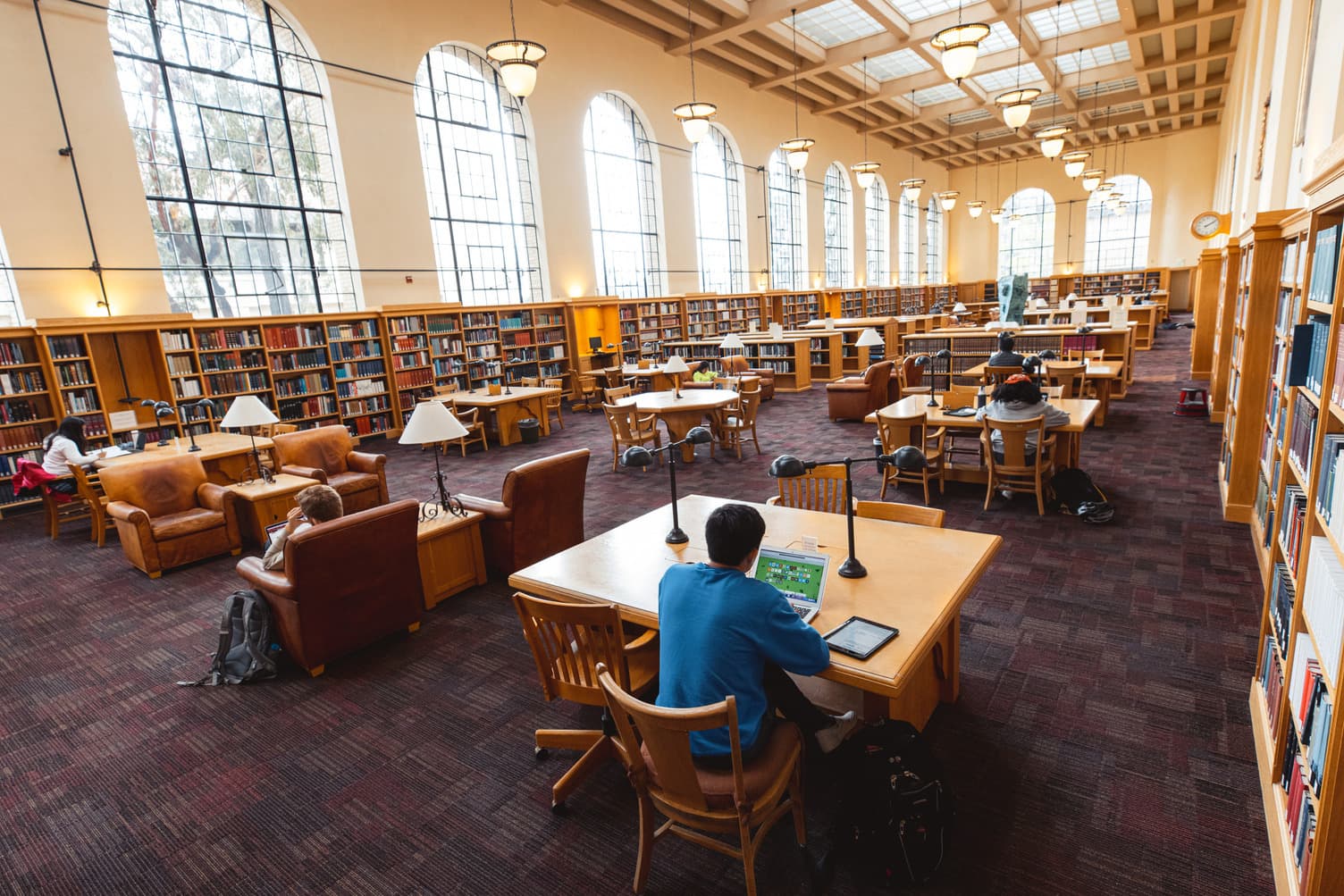 Students are seated, studying at tables in a large open room in a library with many windows.