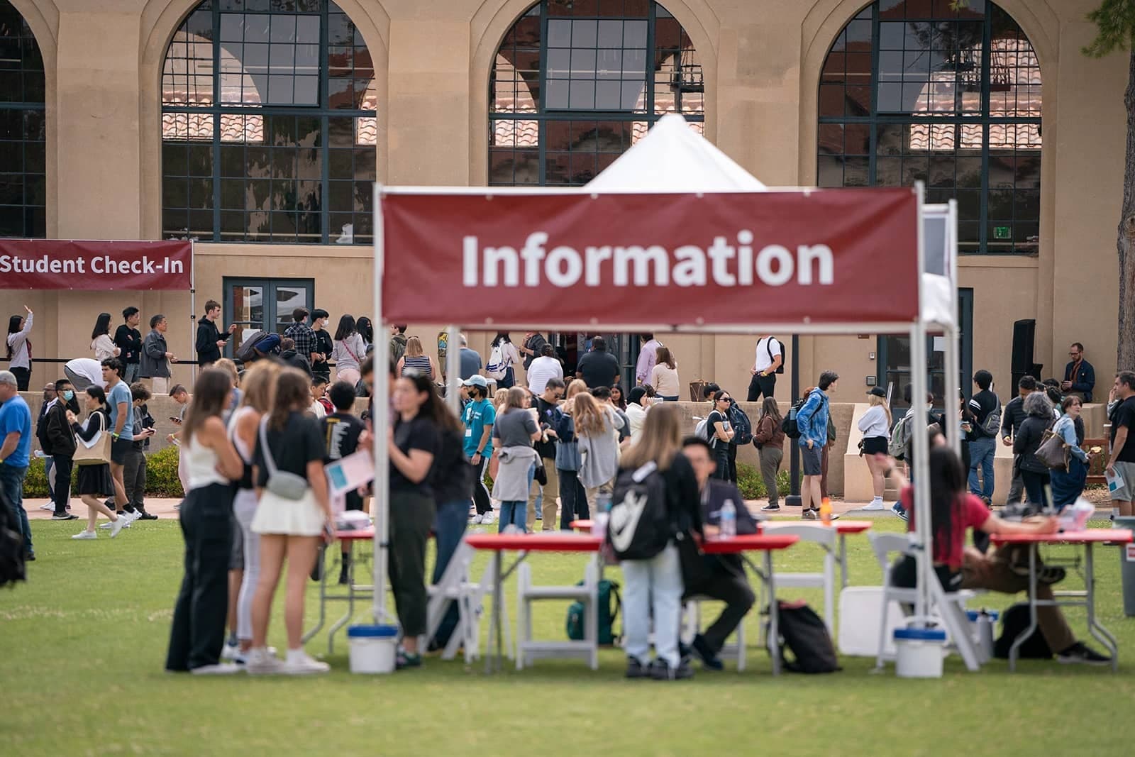 Students stand outside on a large field at an information booth. In the background, other students line up outside a building with arched windows.
