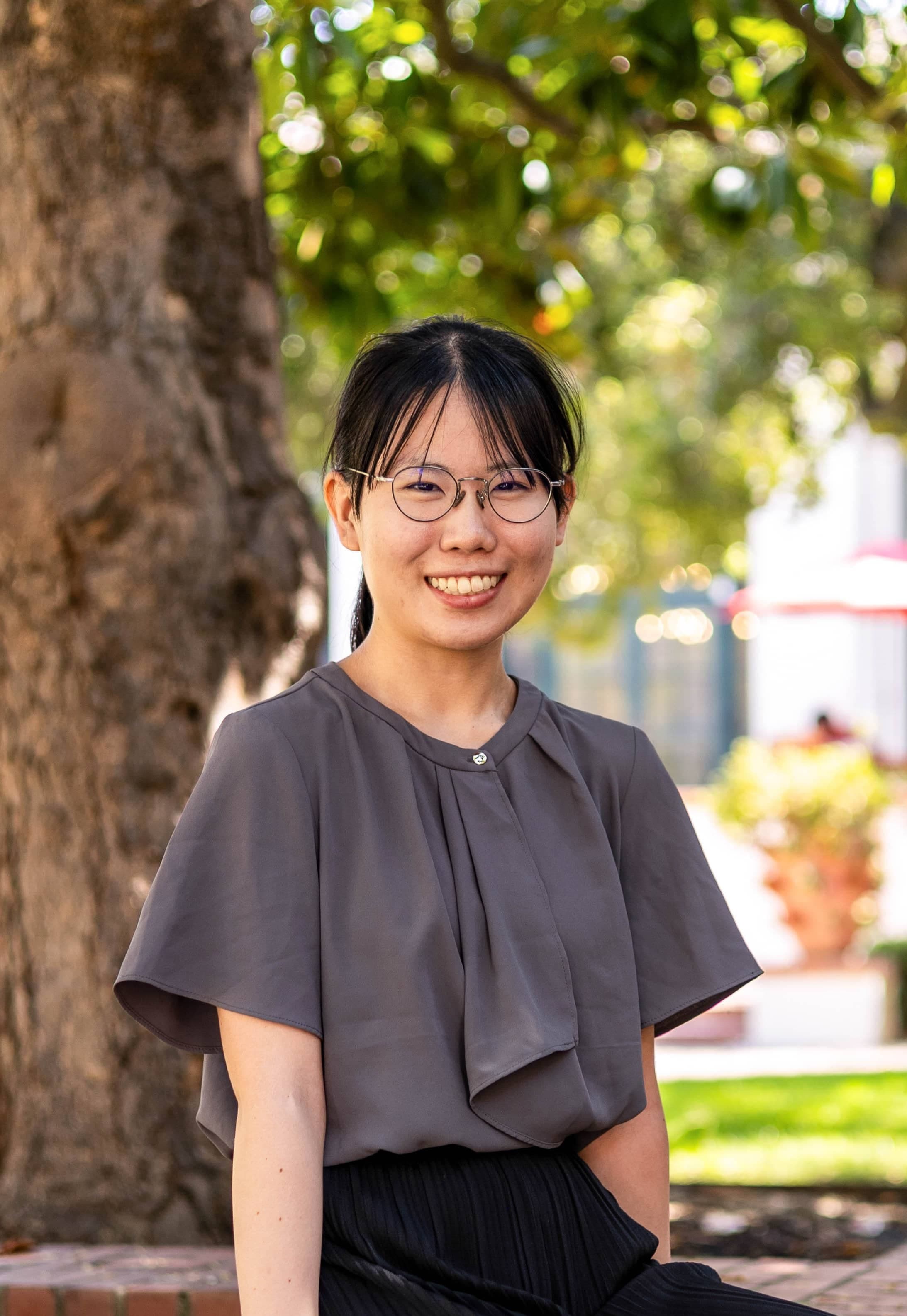 Maki wears a gray top and sits on a wall outside.