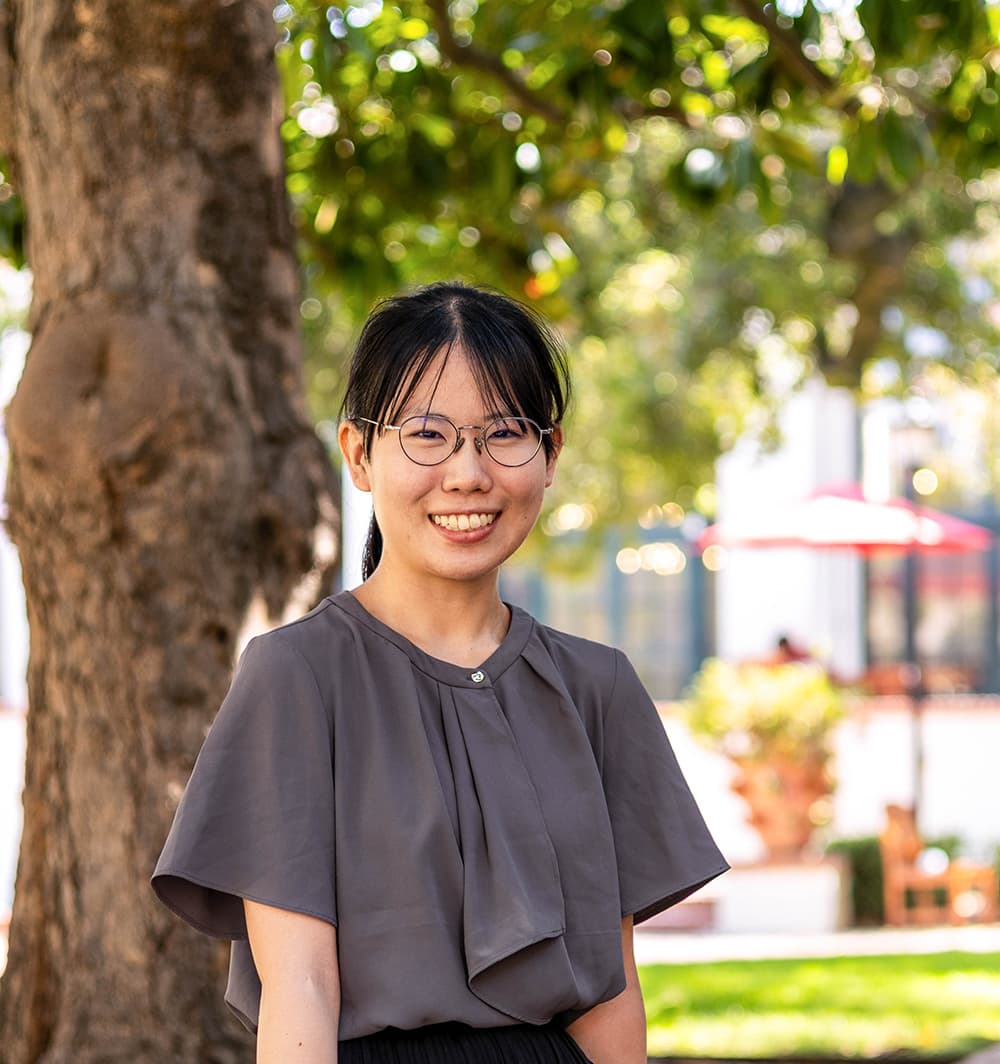 Maki wears a gray top and sits on a wall outside.