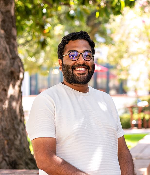Ratna wears a white shirt and sits outside under a tree.